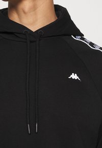 Felpa nera con cappuccio e coulisse, con logo Kappa bianco sul petto e dettagli laterali bianchi a contrasto. Tessuto morbido, vestibilità comoda.