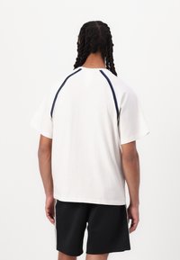 Camiseta blanca de manga corta con mangas raglán que presenta acentos en navy. Textura de algodón suave, corte holgado, vista trasera, combinada con pantalones cortos negros.