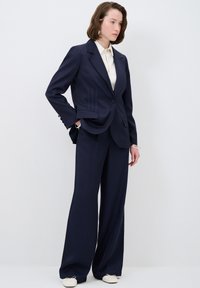 Blazer navy con un solo bottone, con rever tondeggianti e cuciture laterali sottili. Abbinato a pantaloni navy a gamba larga e una camicia color crema.