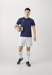 Tumesinise värviga spordisärk, valged püksid, mustad jalgpallijalatsid ja valged sokid, hoides jalgpalli; kujunduses on Puma logoga detailid.