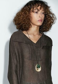 Blouse en lin marron avec col et détails de boutons, associée à un collier pendentif comprenant une pierre verte et des accents dorés.
