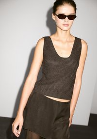 Modelka ve tmavém bezramenném topu a splývavé sukni, s elegantními slunečními brýlemi, stojící na minimalistickém pozadí.