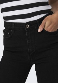 Jeans negros de cintura alta con un bolsillo delantero, cierre de botón metálico y ligera elasticidad. La tela tiene una textura suave y un corte clásico.