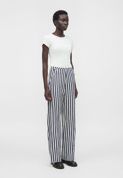 Γυναίκα που φοράει άσπρο τοπ με κοντά μανίκια και ψηλόμεσο παντελόνι σε ναυτικό μπλε και άσπρες κάθετες ρίγες, φαρδιά με μαύρα παπούτσια.