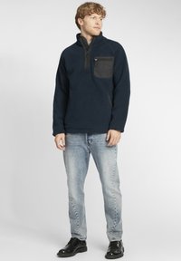 Navy fleece pullover with a quarter-zip collar and black chest pocket; features textured fabric and a relaxed fit. Worn with light jeans and black shoes.