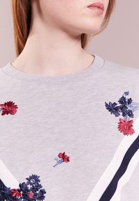 Felpa grigia chiaro con ricami floreali in rosso, blu e rosa, caratterizzata da un collo rotondo e dettagli a righe lungo i lati.