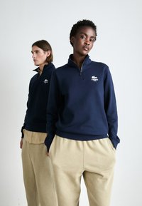 Jūras spēku zipsvārks ar Lacoste logo kreisajā krūtī, kombinēts ar bežām platām biksēm, attēlots uz diviem modeļiem uz neitrāla fona.
