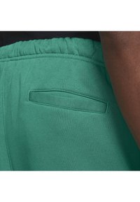 Näruppifrån av gröna byxor med elastisk midja och en horisontell slitsbakficka, bärs av en person med mörk hud och en svart topp.