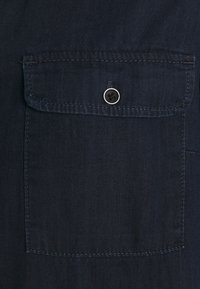 Tkanina ciemnoniebieskiej koszuli z kieszonką na piersi z zapięciem na guzik. Gładka tekstura i wzmocnione szwy wokół kieszonki.