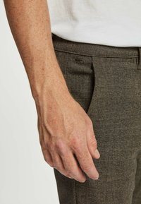 Brūnas šaha raksta bikses, kas izliktas ar roku, balstoties uz auduma, parādot tā tekstūru un dizaina detaļas, tostarp smalku krokainumu.