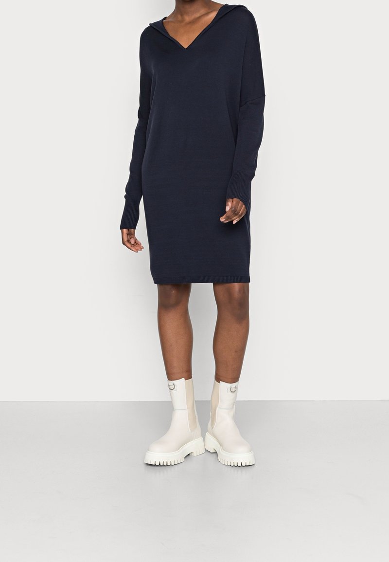 Robe en maille marine avec un col en V et une capuche, dotée de manches longues avec des bas de manches côtelés. Portée avec des bottines blanches à semelle en caoutchouc texturée.