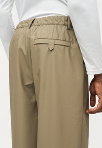 Κοντινό πλάνο ενός ατόμου που φορεί μπεζ παντελόνι με ελαστική μέση και πίσω τσέπη με λουράκι και κουμπί, συνδυασμένο με λευκό μακρυμάνικο μπλουζάκι.