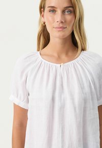 Weißes Leinenbluse mit kurzen Ärmeln, gerafftem Ausschnitt und einem kleinen Knopfdetail in der Mitte, weiche Textur und leichtes Design.