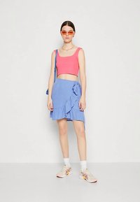 Crop top rose, jupe bleue à volants avec un détail noué, chaussettes blanches et baskets. Accessoires incluant des lunettes de soleil orange et un sac bleu.