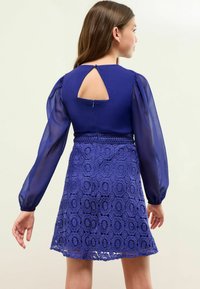 Abito viola con maniche a palloncino trasparenti, dettaglio cut-out sulla schiena e gonna con motivo in pizzo. Dotato di chiusura con cerniera sul retro.