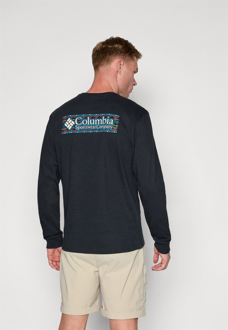 Chemise noire à manches longues en coton, avec un logo graphique coloré au dos, ornée de motifs géométriques et de texte. Associée à un short en khaki.