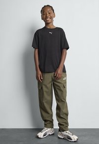 Zwart t-shirt met wit logo, olijfgroene cargobroek met zakken en beige sneakers met zwarte en bordeaux accenten.