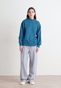 Felpa con cappuccio color teal, dal taglio comodo, abbinata a jeans a gamba larga grigi chiari. L'outfit è completato da sneakers bianche.