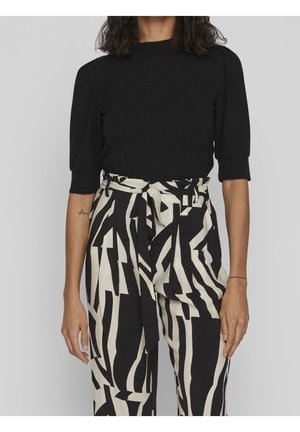 Femme portant un haut noir à manches courtes rentré dans un pantalon taille haute noir et blanc à motif abstrait avec une ceinture en tissu nouée à la taille.