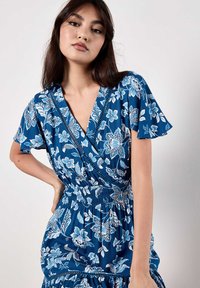 Modrá květinová šaty mají krátké rukávy, zavinovací design, elastický pas a vrstevnatou sukni. Tkanina má hladkou strukturu a živý potisk.