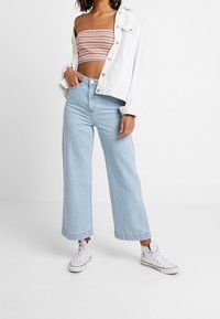 Vrouw in lichtblauwe wijde banden jeans, witte high-top sneakers, een kleurrijke gestreepte tube top, en een witte jas over de schouders gedrapeerd.