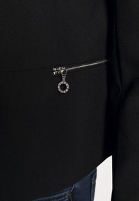 Veste noire en tissu lisse et texturé. Elle présente une fermeture éclair latérale avec un tirette circulaire et des accents de strass. Vue en gros plan sur la zone de la fermeture éclair.