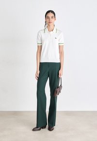 Witte polo met groene accenten, gecombineerd met een donkergroene wijde broek. Model houdt een kleine bruine handtas vast. Eenvoudig, modern ontwerp.