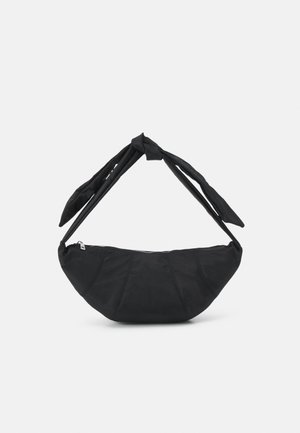 Sort stofhåndtaske med rundet bund og tyk skulderrem bundet i en knude, vist på en ensfarvet lys baggrund.