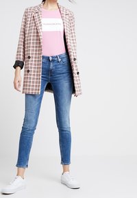 En dubbelknäppt beige och rosa rutmönstrad blazer över en rosa logot-skjorta, kombinerad med slim-fit blå jeans och vita sneakers.