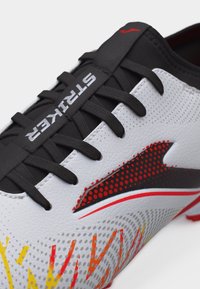 Κοντινό πλάνο ενός λευκού αθλητικού παπουτσιού με μαύρα κορδόνια, κόκκινες και κίτρινες γραφιστικές λεπτομέρειες και το κείμενο "STRIKER" στη γλώσσα του παπουτσιού.