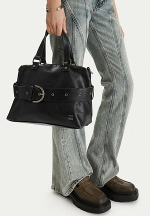 Persona che indossa jeans svasati a lavaggio chiaro e scarpe con plateau marroni, che tiene una borsa nera in pelle con dettaglio di cintura larga.