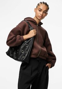 Bruine zip-up hoodie met een losse fit, gecombineerd met een zwarte leren totebag met ronde uitsparingen en zilveren oogjes. Zwarte geplooide broek.