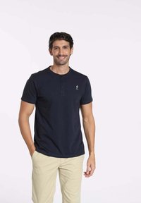 Chemise bleu marine à manches courtes avec un col rond, patte de boutonnage à deux boutons et un petit logo sur la poitrine gauche. Portée avec un pantalon clair.