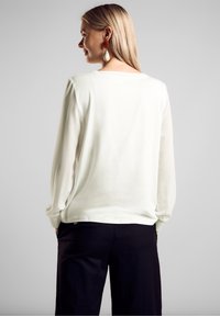 Blusa bianca a maniche lunghe, texture liscia, scollatura rotonda, vestibilità comoda, abbinata a pantaloni neri a gamba larga. La vista posteriore mette in mostra la semplicità del design.