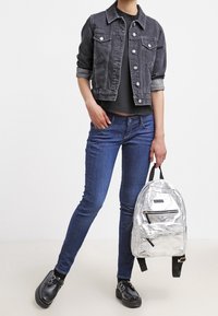 Chaqueta de mezclilla gris oscuro, top negro ajustado, jeans ajustados azules y mochila plateada metálica con cremalleras. Zapatos negros con detalle perforado.