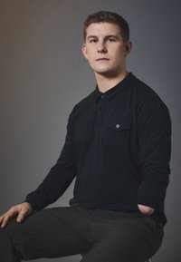 Granatowa koszulka polo z długim rękawem, z zapięciem na guziki przy kołnierzy oraz kieszonką na piersi, w połączeniu z szarymi spodniami w wzory, siedząca na tle w neutralnych kolorach.