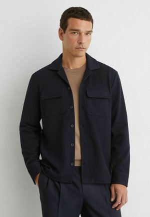 Mann trägt eine marineblaue Jacke mit Knöpfen und Brusttaschen über einem beigen Hemd, steht vor einer weißen Wand.