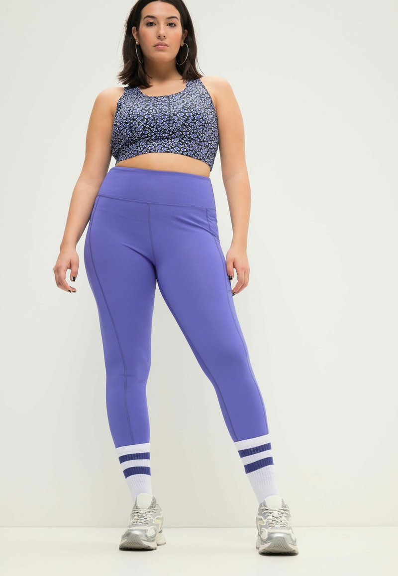 Paarse leggings met zijnaad en hoge tailleband, gecombineerd met een aansluitende zwarte en blauw patroon crop top, gedragen met witte sokken met blauwe strepen.