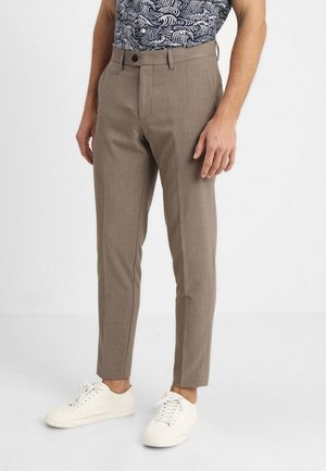 Homme portant un pantalon beige slim, des baskets blanches et une chemise à manches courtes bleue à motifs, debout devant un fond uni.