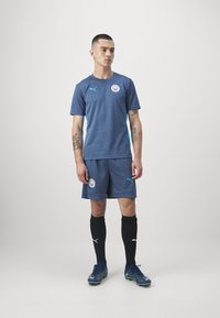 Jūras spēku sporta komplekts ar īsām piedurknēm un šortiem, ar gaiši ziliem akcentiem un komandas logo. Melni zeķbikses un rakstaini sporta apavi noslēdz izskatu.