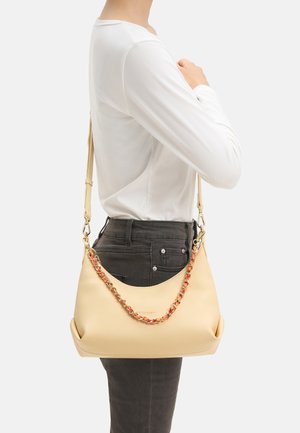 Person wearing a white long-sleeve shirt and dark pants holding a beige handbag with a gold and red chain strap over their shoulder.