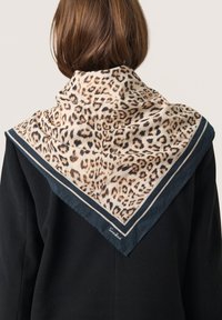 Løveprint tørklæde lavet af blødt stof; har en mørk trim med et mønster i creme og brune nuancer. Båret om halsen over en sort frakke.