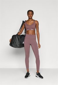 Sportbeha en legging in gedempt mauve, met een Nike-logo op de band. Zwarte duffeltas en sneakers maken de sportieve outfit compleet.