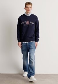 Tengerész navy pulóver "GANT 1949" logóval, világoskék farmernadrággal párosítva. A modellen fehér sportcipők vannak, ami egy laza, kényelmes stílust tükröz.