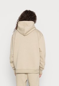 Béžová mikina s velkou kapucí, spadlými švy na ramenou a žebrovanými manžetami. Vyrobena z měkké látky pro pohodlí. Jednoduchý, minimalistický design.