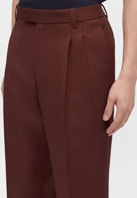 Personne portant un pantalon marron taille haute avec passants pour ceinture et plis devant, main reposant à côté de la cuisse droite.