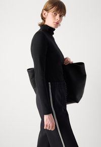 Un pull à col roulé noir à manches longues associé à un pantalon sombre avec des rayures blanches, tenant un grand sac fourre-tout en cuir noir. Design simple et moderne.