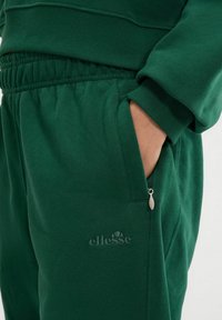 Pantalones de chándal verdes fabricados en tejido suave con cintura elástica, bolsillo lateral con cierre de cremallera y logotipo en relieve en la parte frontal.