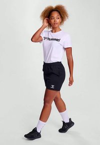 Λευκό T-shirt με μαύρο λογότυπο, μαύρα σορτς και μαύρα αθλητικά παπούτσια. Το μοντέλο φοράει άσπρες κάλτσες, αναδεικνύοντας ένα casual αθλητικό στυλ. Σγουρά μαλλιά.