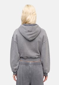 Grijze cropped hoodie met een grote capuchon, geribbelde manchetten en tailleband. Bevat geborduurde branding op de bovenrug. Zachte stoftextuur.
